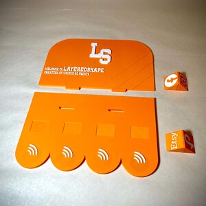 May include: Orange layered shape display pieces with white text that reads "LS" and "WELCOME TO LAYEREDSHAPE CREATORS OF UNIQUE 3D PRINTS". Two small orange and white cubes with Etsy and other logos are also present.