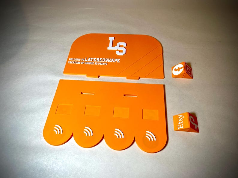 May include: Orange layered shape display pieces with white text that reads "LS" and "WELCOME TO LAYEREDSHAPE CREATORS OF UNIQUE 3D PRINTS". Two small orange and white cubes with Etsy and other logos are also present.
