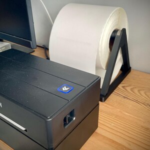 May include: A black label printer with the brand name "MUNBYN" is on a wooden surface. A large roll of white labels sits in a black holder. The printer has a blue power button with an arrow symbol.