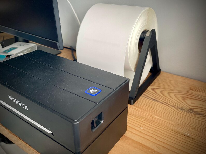 May include: A black label printer with the brand name "MUNBYN" is on a wooden surface. A large roll of white labels sits in a black holder. The printer has a blue power button with an arrow symbol.