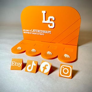 May include: Orange display stand with white text and social media icons. The stand features the text "WELCOME TO LAYEREDSHAPE CREATORS OF UNIQUE 3D PRINTS" and the initials "LS". Individual blocks display Etsy, TikTok, Facebook, and Instagram logos.