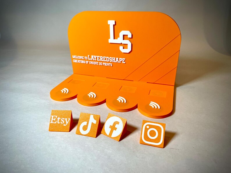 May include: Orange display stand with white text and social media icons. The stand features the text "WELCOME TO LAYEREDSHAPE CREATORS OF UNIQUE 3D PRINTS" and the initials "LS". Individual blocks display Etsy, TikTok, Facebook, and Instagram logos.