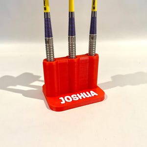 Op de afbeelding: Een rode 3D-geprinte darthouder met de naam "JOSHUA" in witte letters. Drie darts met gele, paarse en zilveren accenten zijn erin geplaatst. De houder staat op een witte ondergrond.