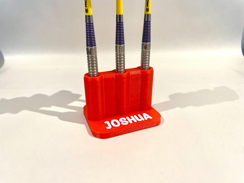 Op de afbeelding: Een rode 3D-geprinte darthouder met de naam "JOSHUA" in witte letters. Drie darts met gele, paarse en zilveren accenten zijn erin geplaatst. De houder staat op een witte ondergrond.