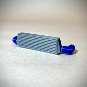 May include: A grey intercooler with blue silicone hoses. The intercooler has a rectangular shape with horizontal fins. The blue hoses are curved and connect to each end of the intercooler. The object is a miniature model.