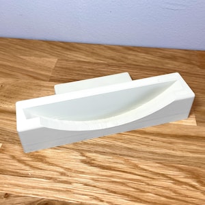 May include: White plastic phone stand with a curved, indented area for holding a phone.