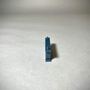 May include: A small, rectangular, teal-colored object with two small, cylindrical protrusions on top and a small, circular detail on the front. The object is standing upright against a plain, light-colored background.