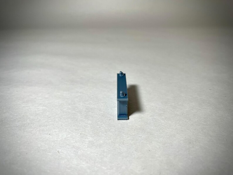 May include: A small, rectangular, teal-colored object with two small, cylindrical protrusions on top and a small, circular detail on the front. The object is standing upright against a plain, light-colored background.
