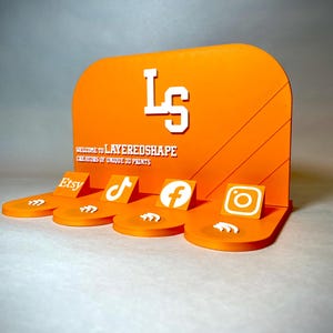May include: An orange display stand with the text "LS" and "LAYEREDSHAPE" in white. The stand features platforms with Etsy, TikTok, Facebook, and Instagram logos. The design is made of 3D printed material.