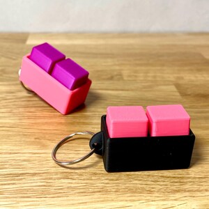 May include: Two keychains with two small square blocks attached. The keychain on the left has pink and purple blocks and a pink holder. The keychain on the right has pink blocks and a black holder.