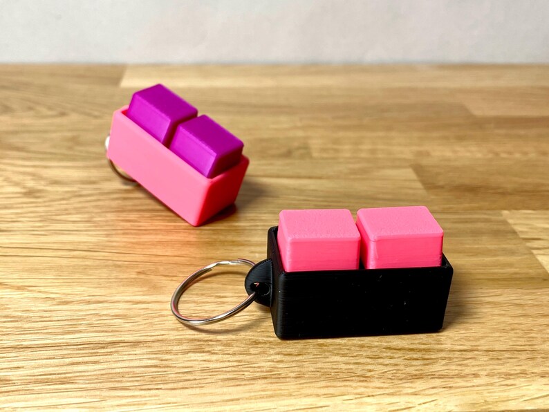 May include: Two keychains with two small square blocks attached. The keychain on the left has pink and purple blocks and a pink holder. The keychain on the right has pink blocks and a black holder.