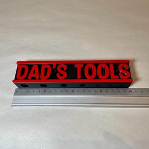 May include: A black and red tool organizer with the text "DAD'S TOOLS" in red letters. The organizer has several holes for tools and is approximately 10 inches long. A metal ruler is placed next to the organizer.