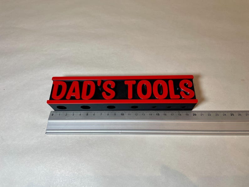 May include: A black and red tool organizer with the text "DAD'S TOOLS" in red letters. The organizer has several holes for tools and is approximately 10 inches long. A metal ruler is placed next to the organizer.