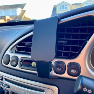 May include: A black car phone mount attached to the air vent in a car's dashboard. The mount is made of plastic and has a spring-loaded clamp to hold a phone.