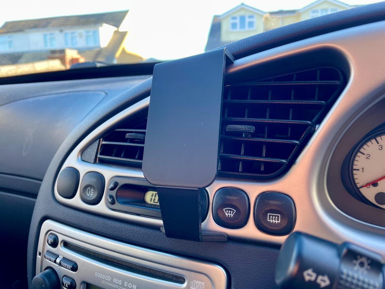 May include: A black car phone mount attached to the air vent in a car's dashboard. The mount is made of plastic and has a spring-loaded clamp to hold a phone.