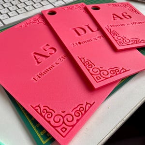 May include: Three rectangular pink templates with rounded corner cutouts and circular holes. The templates are labeled A5 (148mm x 210mm), DL (210mm x 99mm), and A6 (148mm x 105mm). Each piece features embossed decorative corner scrollwork patterns.