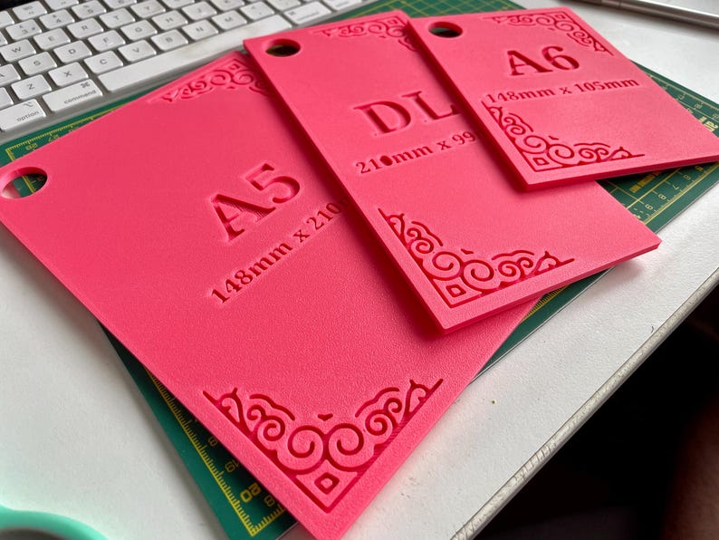 May include: Three rectangular pink templates with rounded corner cutouts and circular holes. The templates are labeled A5 (148mm x 210mm), DL (210mm x 99mm), and A6 (148mm x 105mm). Each piece features embossed decorative corner scrollwork patterns.