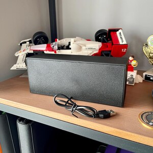 May include: A black rectangular speaker with a USB cable sits on a wooden shelf. A Lego race car, a trophy, and a medal are also on the shelf. The speaker is a simple, modern design.