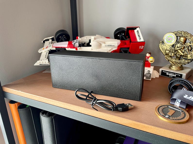 May include: A black rectangular speaker with a USB cable sits on a wooden shelf. A Lego race car, a trophy, and a medal are also on the shelf. The speaker is a simple, modern design.