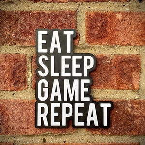 May include: Black sign with white text that reads "EAT SLEEP GAME REPEAT" against a red brick wall. The sign is a single piece with the words stacked vertically. The brick wall has a rough texture.