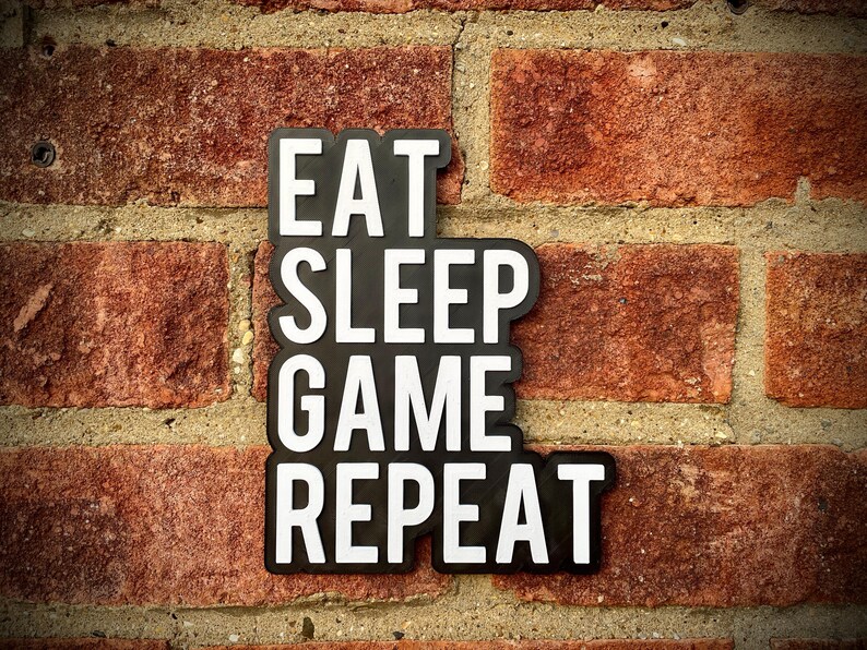 May include: Black sign with white text that reads "EAT SLEEP GAME REPEAT" against a red brick wall. The sign is a single piece with the words stacked vertically. The brick wall has a rough texture.