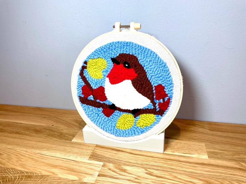 May include: A hand-stitched embroidery hoop with a blue background and a red-breasted robin perched on a branch with yellow leaves. The robin is brown, white, and red.