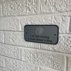 May include: A gray metal sign with rounded corners is mounted on a white brick wall. The sign has the text "I'm out seeing a man about a dog." A dog graphic is in the upper center.