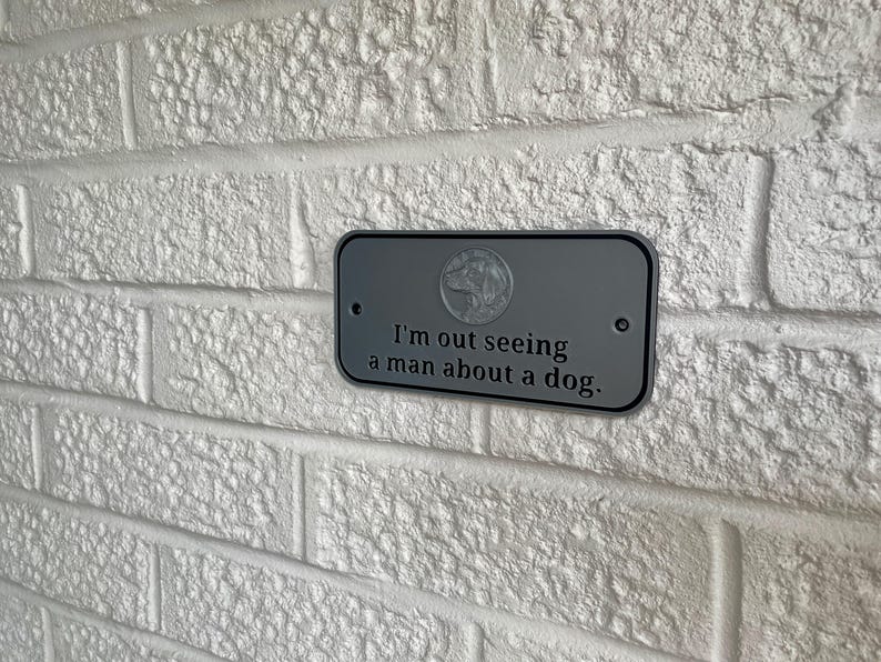 May include: A gray metal sign with rounded corners is mounted on a white brick wall. The sign has the text "I'm out seeing a man about a dog." A dog graphic is in the upper center.