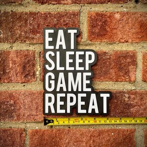 May include: Black and white sign with the text "EAT SLEEP GAME REPEAT" against a red brick wall. A yellow measuring tape is placed below the sign, showing measurements in inches.