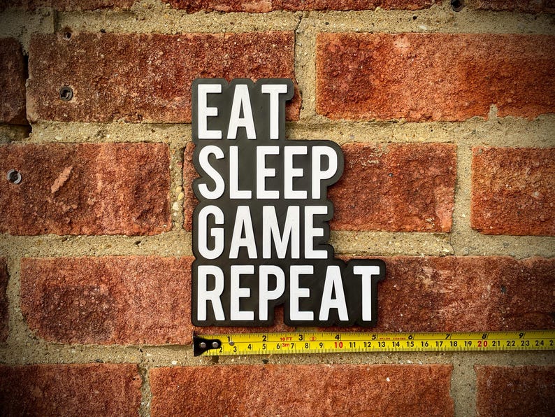 May include: Black and white sign with the text "EAT SLEEP GAME REPEAT" against a red brick wall. A yellow measuring tape is placed below the sign, showing measurements in inches.