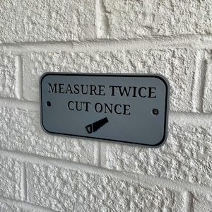 May include: A rectangular, gray metal sign with rounded corners is mounted on a white brick wall. The sign reads "MEASURE TWICE CUT ONCE" in black text, with a saw graphic below. Two small screws secure the sign.
