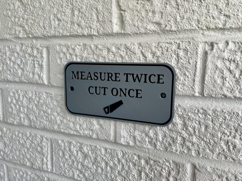 May include: A rectangular, gray metal sign with rounded corners is mounted on a white brick wall. The sign reads "MEASURE TWICE CUT ONCE" in black text, with a saw graphic below. Two small screws secure the sign.