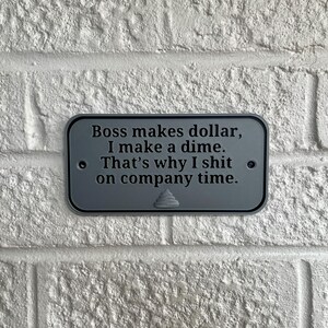 May include: A rectangular, grey metal sign with rounded corners is mounted on a white brick wall. The sign has black text that reads, "Boss makes dollar, I make a dime. That's why I shit on company time."