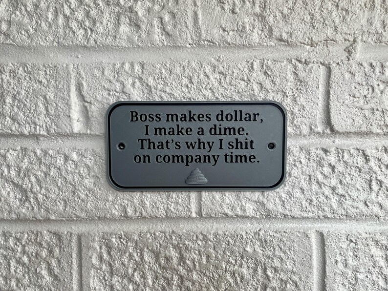 May include: A rectangular, grey metal sign with rounded corners is mounted on a white brick wall. The sign has black text that reads, "Boss makes dollar, I make a dime. That's why I shit on company time."