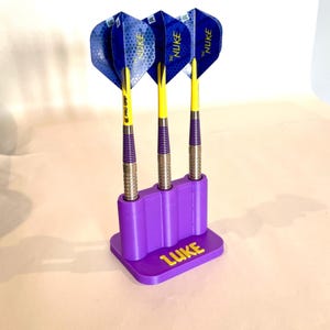 Op de afbeelding: Drie darts met blauwe en gele flights worden getoond in een paarse houder. De darts hebben zilveren barrels, gele shafts en paarse grips. De houder heeft de naam "LUKE" in geel.