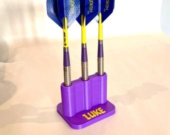 May include: Three darts with blue and yellow flights are displayed in a purple holder. The darts have silver barrels, yellow shafts, and purple grips. The holder has the name "LUKE" in yellow.