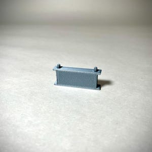 May include: A small, gray, rectangular object with two vertical supports and two small cylindrical protrusions on top. The object appears to be made of a textured material and is set against a neutral background.