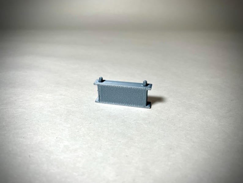 May include: A small, gray, rectangular object with two vertical supports and two small cylindrical protrusions on top. The object appears to be made of a textured material and is set against a neutral background.
