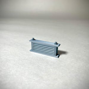 May include: A small, gray, rectangular object with horizontal bars and decorative edges. The object has two small, cylindrical protrusions on top. The item is likely a miniature architectural element or decorative piece.