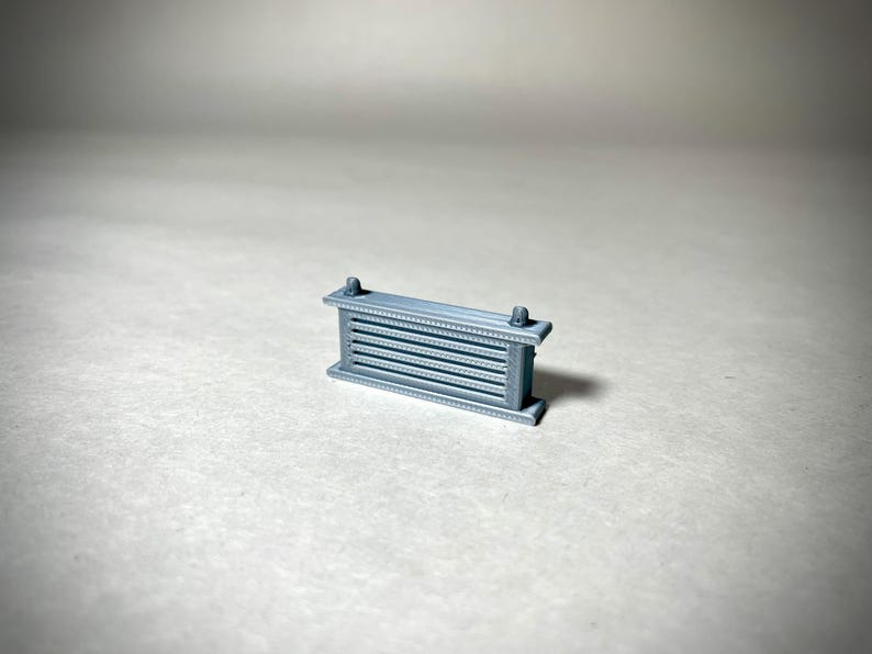 May include: A small, gray, rectangular object with horizontal bars and decorative edges. The object has two small, cylindrical protrusions on top. The item is likely a miniature architectural element or decorative piece.