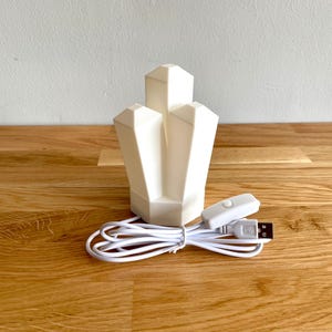 Pu&ograve; includere: Una luce notturna bianca a forma di cristallo con un cavo USB e un interruttore bianco. La luce &egrave; su una superficie di legno.