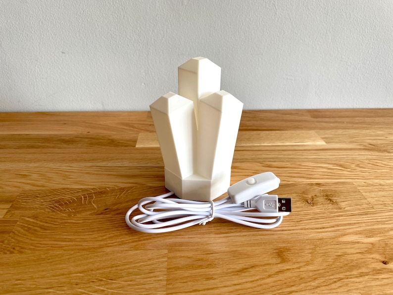 Pu&ograve; includere: Una luce notturna bianca a forma di cristallo con un cavo USB e un interruttore bianco. La luce &egrave; su una superficie di legno.