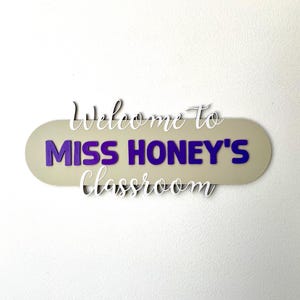 K&ouml;nnte beinhalten: Ein Klassenzimmerschild mit dem Text "Welcome to MISS HONEY'S Classroom". Die Worte "MISS HONEY'S" sind in lila Blockbuchstaben auf einem beigen Oval, die anderen Worte in wei&szlig;er Schrift.