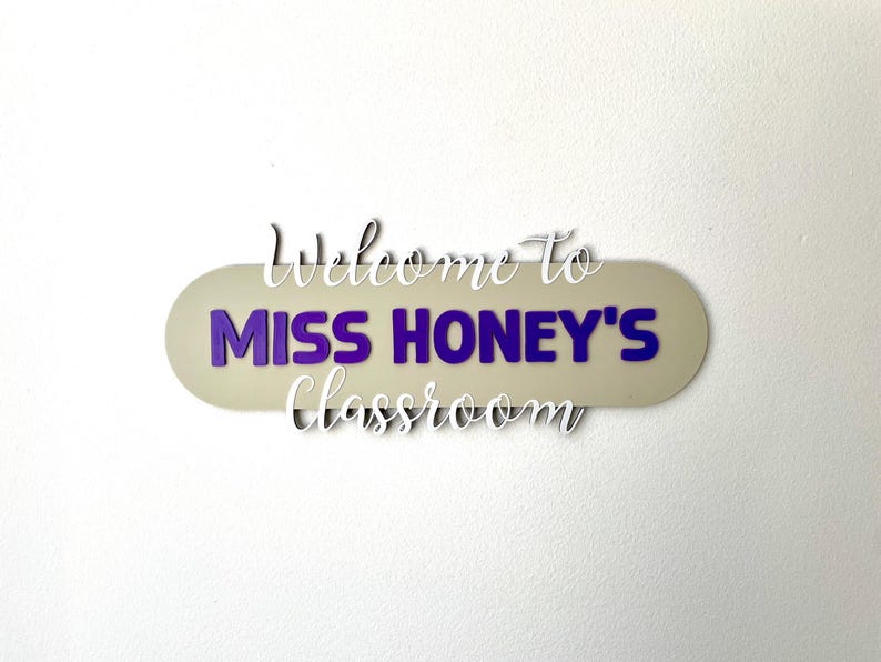 K&ouml;nnte beinhalten: Ein Klassenzimmerschild mit dem Text "Welcome to MISS HONEY'S Classroom". Die Worte "MISS HONEY'S" sind in lila Blockbuchstaben auf einem beigen Oval, die anderen Worte in wei&szlig;er Schrift.