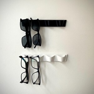 May include: Two wall-mounted eyewear organizers, one black and one white, each holding two pairs of glasses. The top organizer is black and has a straight design, while the bottom one is white with a wavy design.