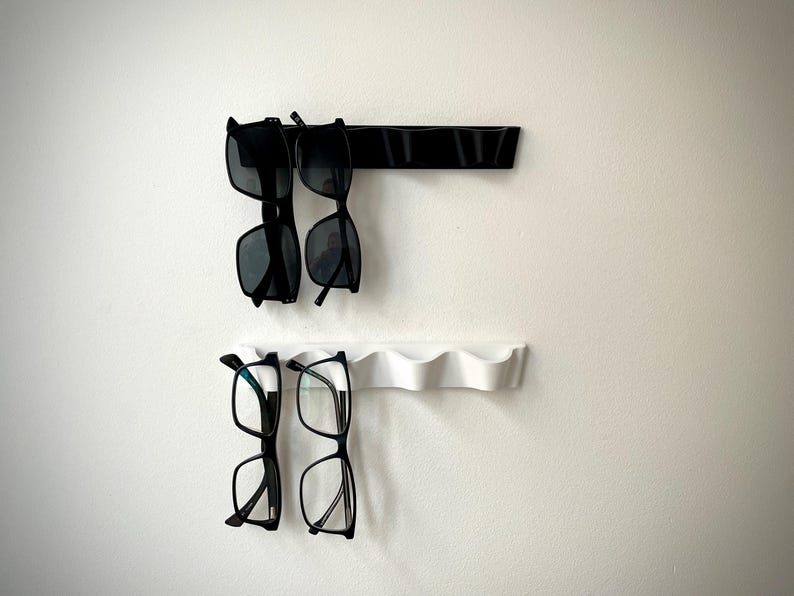 May include: Two wall-mounted eyewear organizers, one black and one white, each holding two pairs of glasses. The top organizer is black and has a straight design, while the bottom one is white with a wavy design.