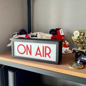 May include: A black rectangular sign with the words "ON AIR" in red letters. The sign is on a wooden shelf with a toy race car, a trophy, and other items.