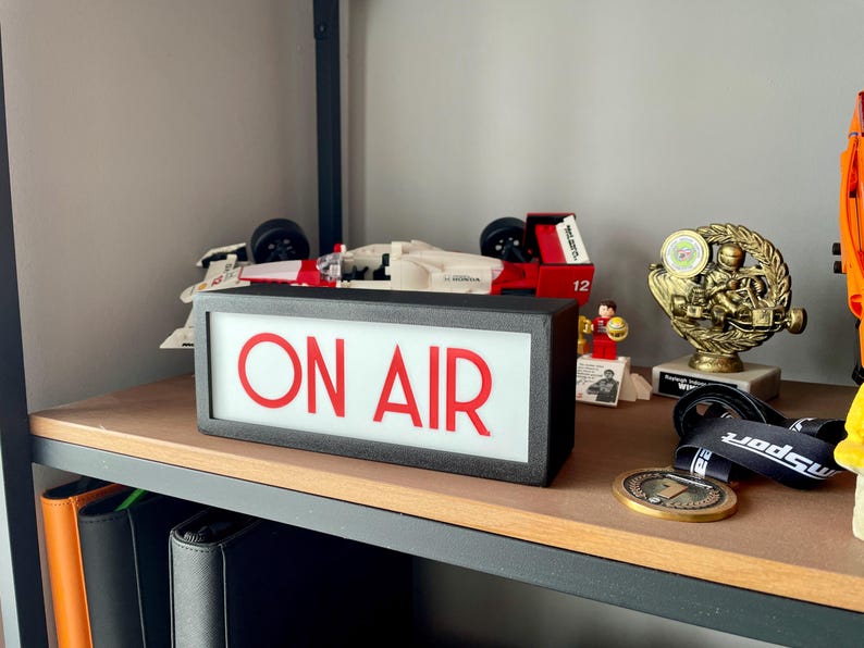 May include: A black rectangular sign with the words "ON AIR" in red letters. The sign is on a wooden shelf with a toy race car, a trophy, and other items.