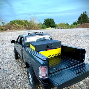 May include: Dark green toy pickup truck with a black toolbox and a yellow toolbox in the bed. The truck has a roof rack and the tailgate is down. The truck is parked on a gravel surface.