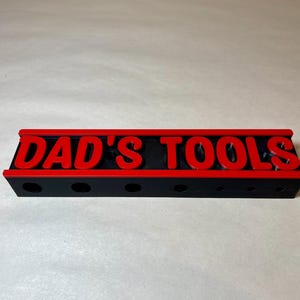 May include: A black tool organizer with the text "DAD'S TOOLS" in red. The organizer has a red border and several holes for holding tools. The item is made of plastic and is designed to hold tools.